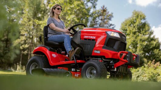 A woman in a gray t-shirt, jeans, and work books sitting on a Craftsman riding mower with trees in the background