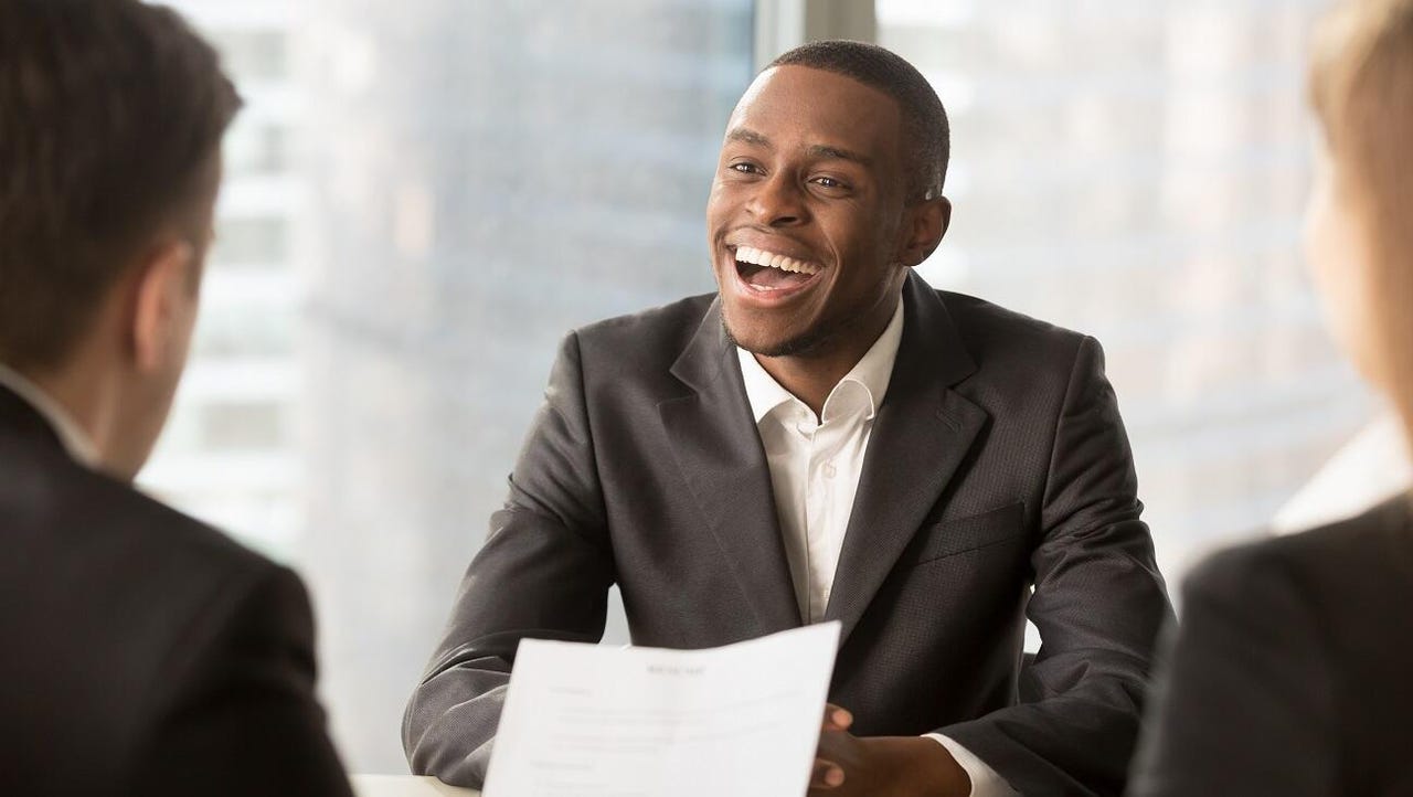 A Black man in business attire smiles after getting hired. He is seated across from two interviewers who face him.