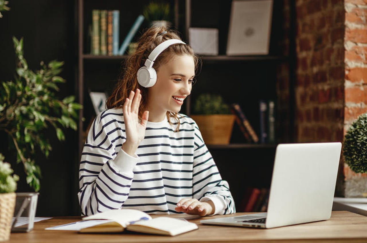 Cheerful woman in headphones greeting friend while talking on laptop at home