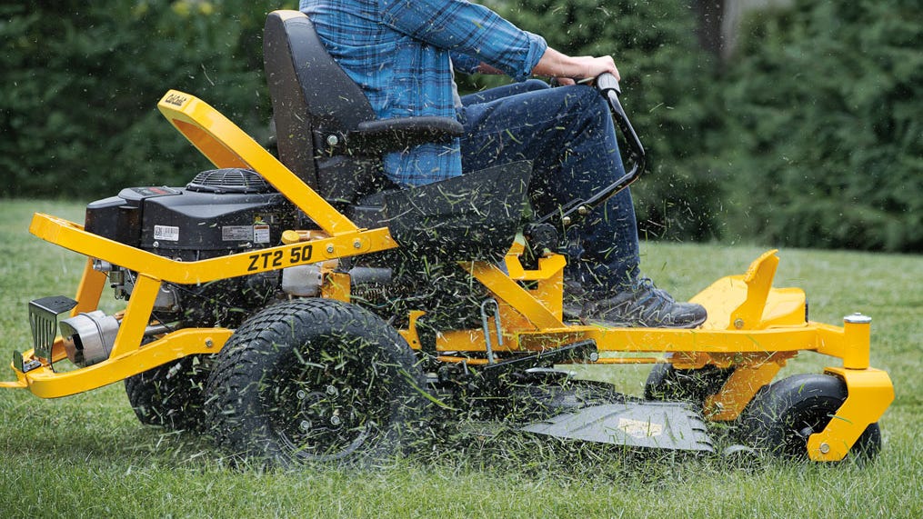 Partial shot of someone in work clothes using a Cub Cadet zero turn mower to cut grass. Grass clippings are flying out of the discharge chute of the cutting deck.