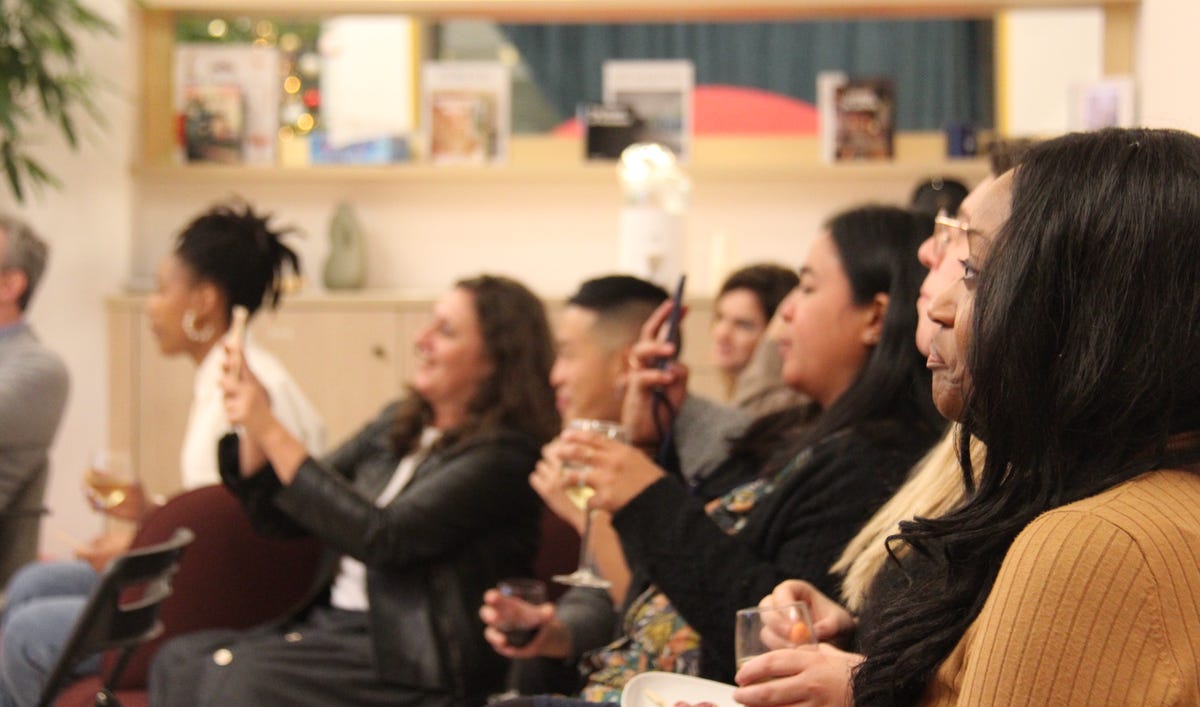 a group of smiling women and men gathered around a warmly-lit room holding drinks and taking photos on their smartphones