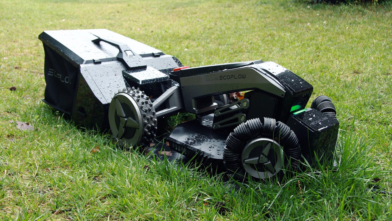 A Husqvarna robot lawn mower cutting grass while a person tends to a flower bed in the background
