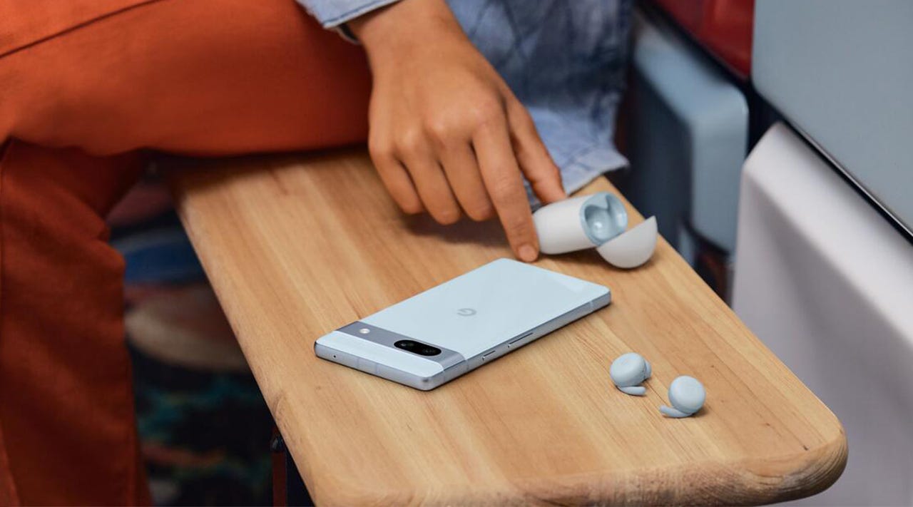 Close-up of a Google Pixel 7a smartphone on a bench as a young woman uses a pair of Google Pixel A-Series earbuds