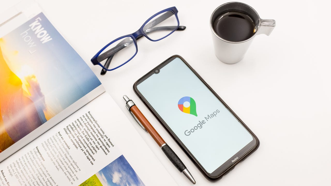 Google Maps logo on a smartphone next to a magazine, a pen, a pair of glasses, and a cup of coffee